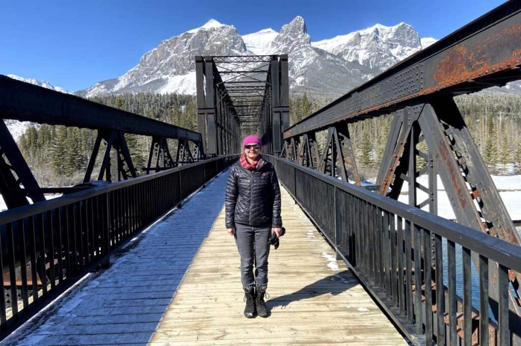 A woman stands on canmore engine bridge on a sunny winters day with snow-capped mountains in the background