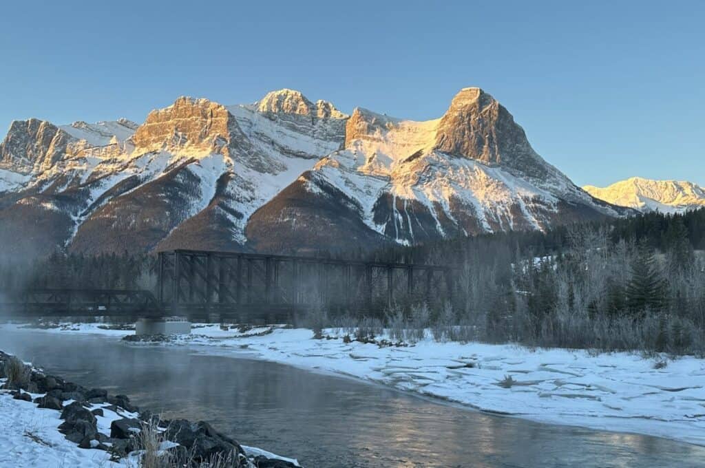 An icy bow river runs beneath the canmore engine bridge at sunrise in the background ha ling peak is lit by the sun